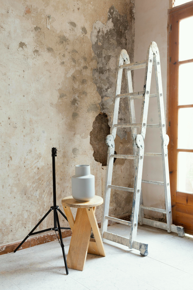 A still-life setup: a grey vase on a wooden stool, ladders against a peeling wall, tripod and a window.