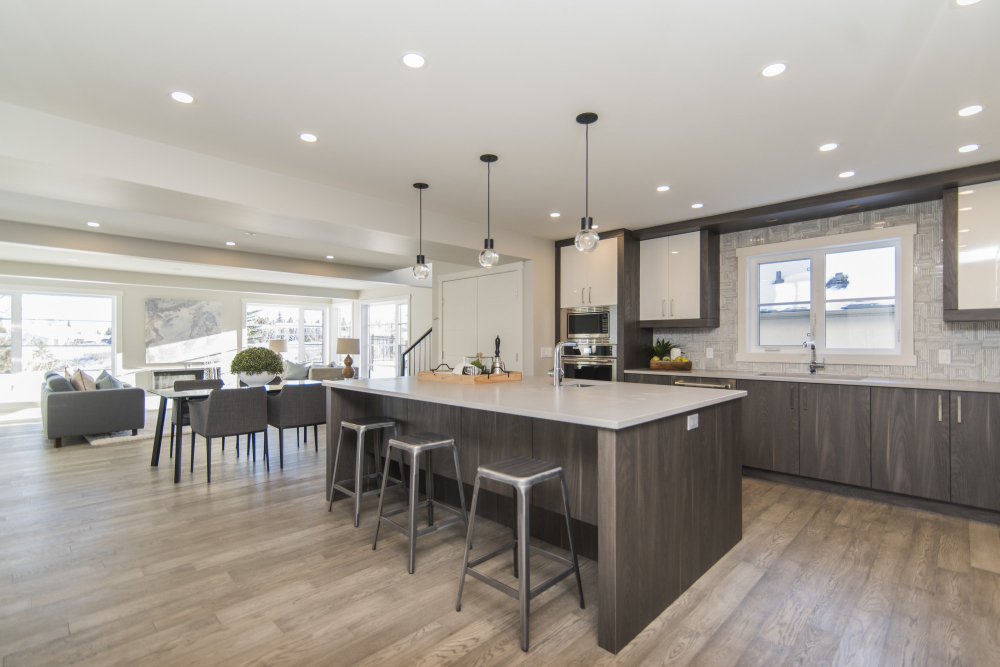 Modern kitchen featuring an island with stools, pendant lights, wooden cabinets, and a dining area. Natural light fills the room.