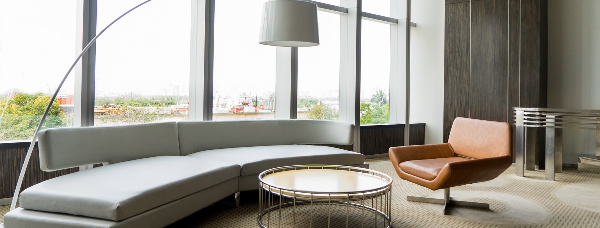 Modern living room with a curved white sofa, round coffee table, leather armchair, and floor lamp near large windows with a city view.